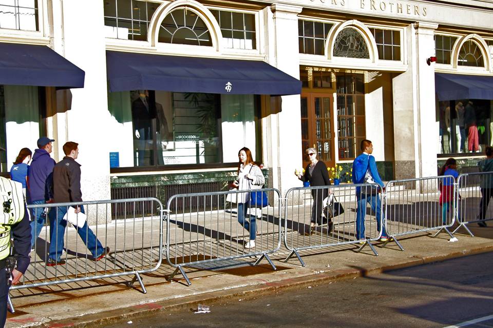 Many people are walking along the commercial street that crowd control barrier is placed.