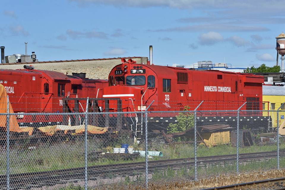 Two locomotives are stopped along the railway tracks surrounded by galvanized chain link fence.