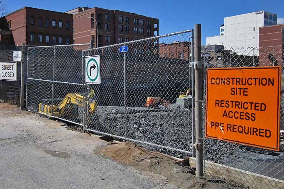 A digger is working in the construction site enclosed by temporary chain link fence with three warning signs.